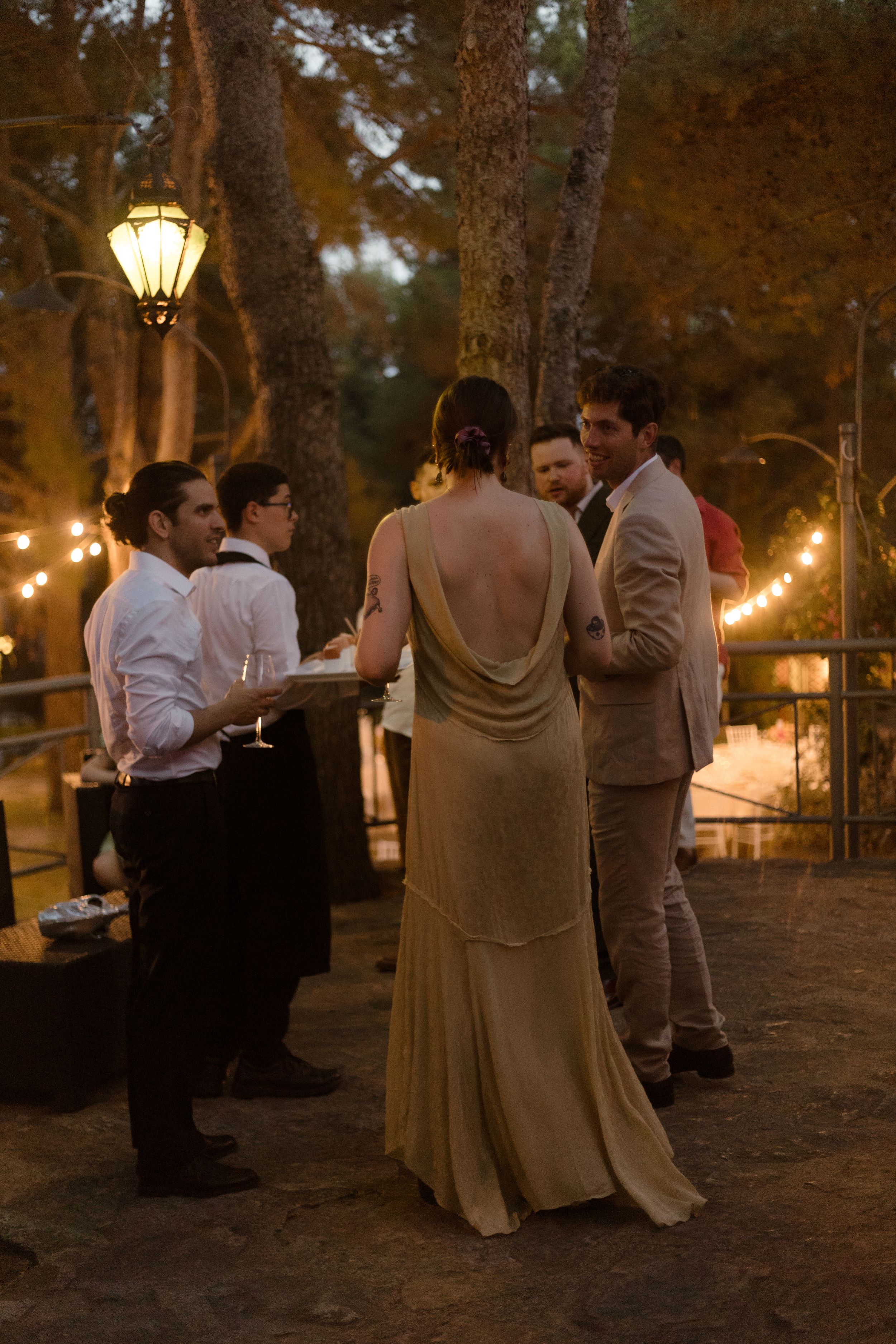An evening outdoor wedding reception with string lights and vintage lanterns illuminating guests mingling under tall trees.