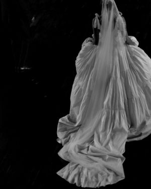 A dramatic black and white photograph of a flowing white wedding dress with a long train and veil against a dark background.