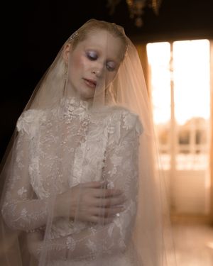 A bride in a delicate lace wedding dress and veil stands in soft window light with eyes closed in a dreamy pose.