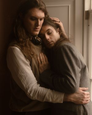 A tender, intimate moment shows two people embracing in warm light by a doorway, wearing vintage-style clothing.