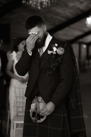 An emotional moment at a Scottish wedding as a groom in traditional tartan kilt and formal jacket wipes his eyes.