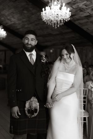 A black and white wedding photo showing a couple beneath a crystal chandelier, the groom in a kilt and the bride in white.