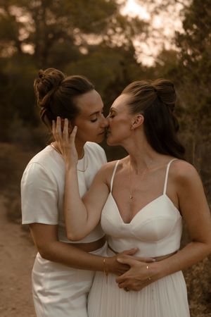 A romantic couple in white outfits share an intimate kiss at sunset on a dirt path surrounded by trees.