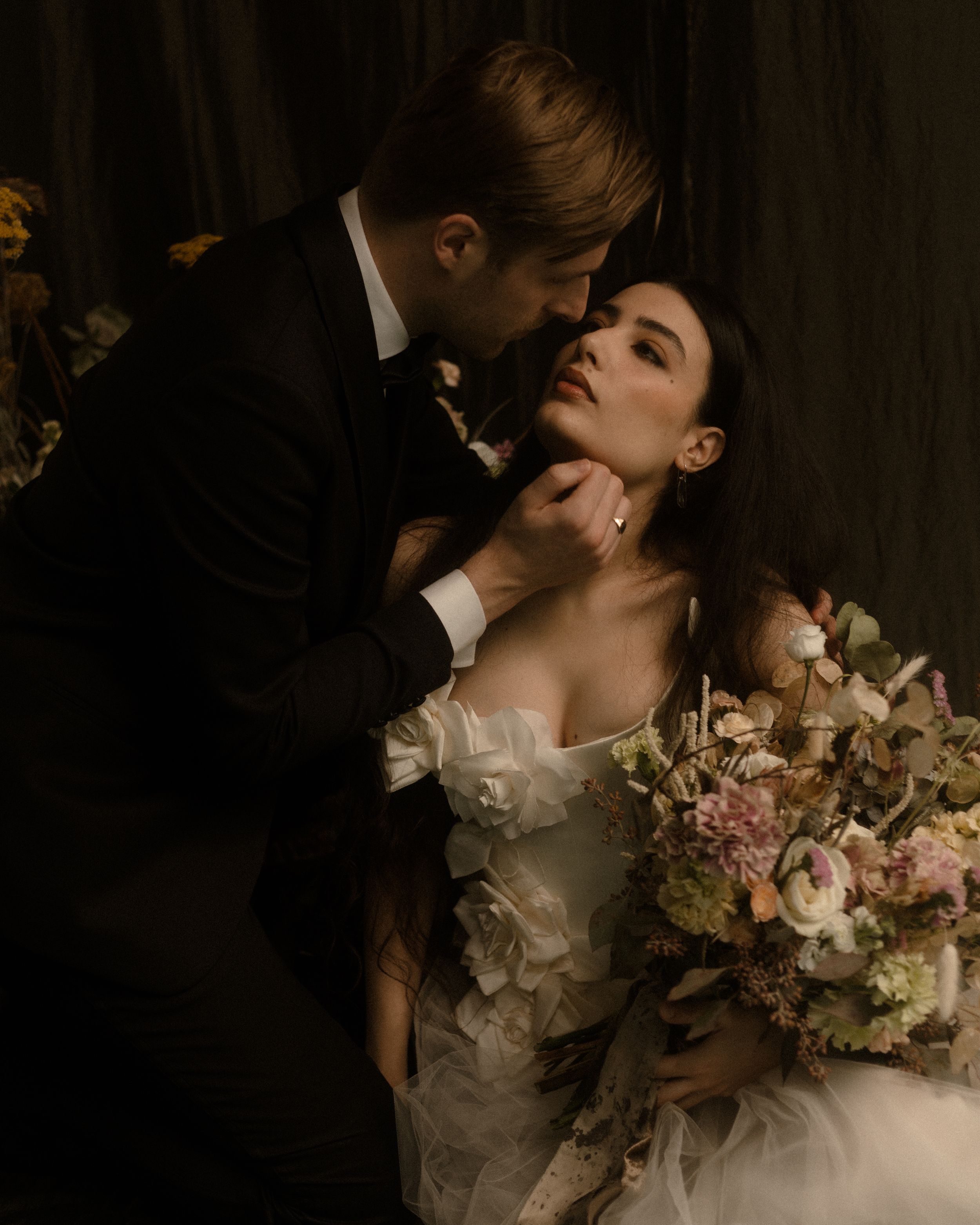 A romantic wedding couple embraces in a moody dark setting with the bride holding a soft pink and white floral bouquet.
