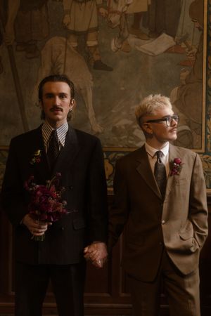 Two well-dressed men in vintage suits stand in an elegant room with wall murals, one holding purple flowers.
