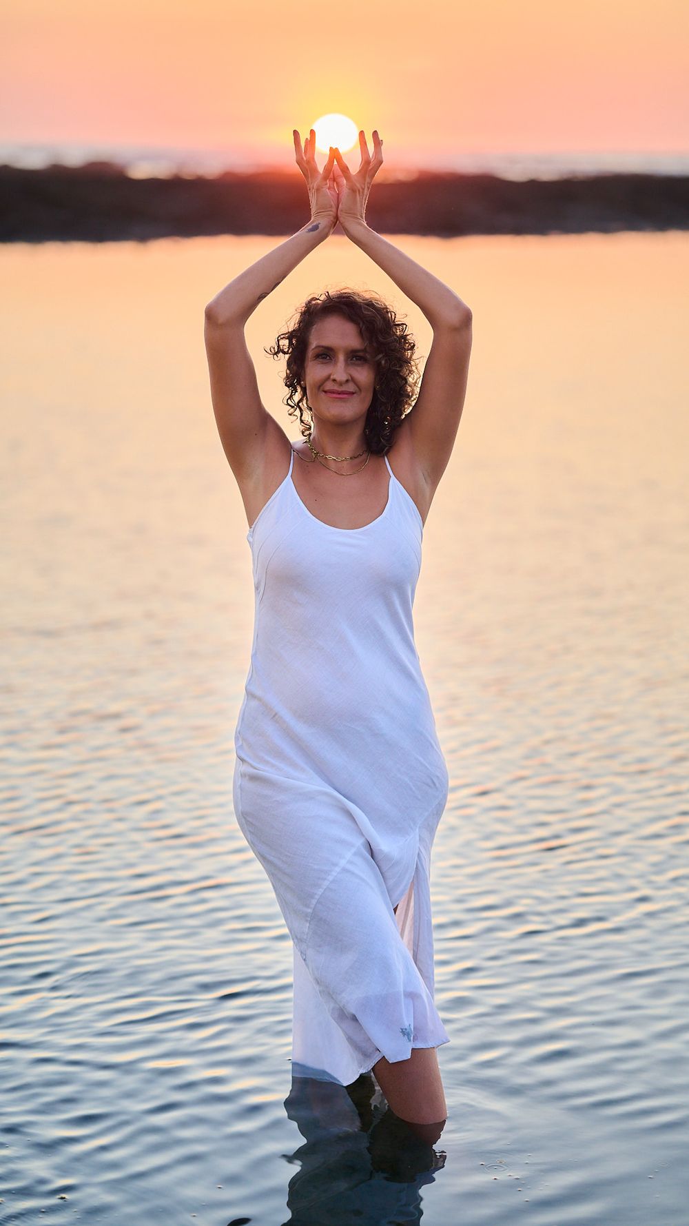A person wearing a white tank top poses playfully by a tranquil lake at sunset with arms raised overhead.