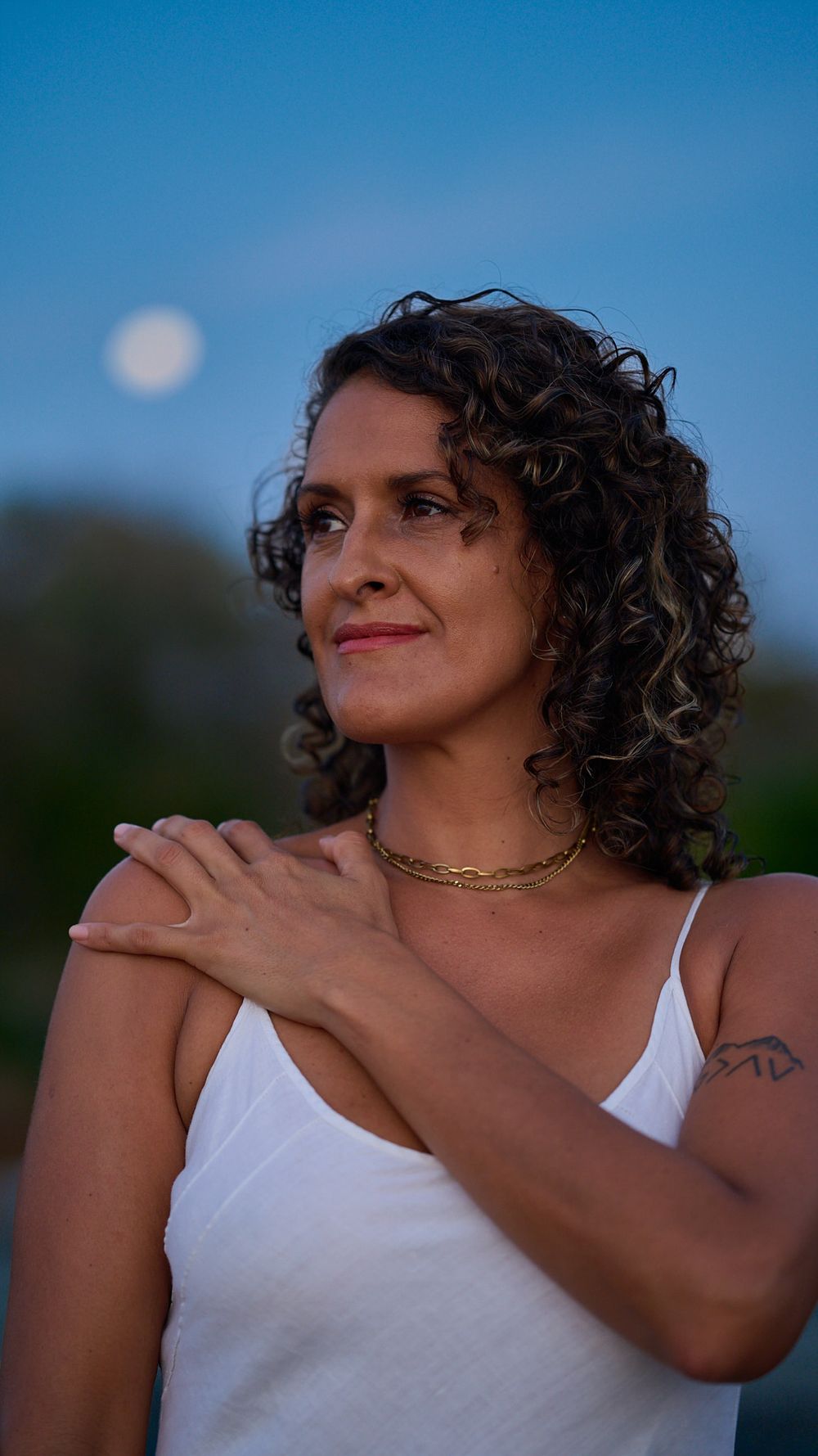 A person with curly brown hair wearing a white tank top poses outdoors at dusk with the moon visible in the blue sky.
