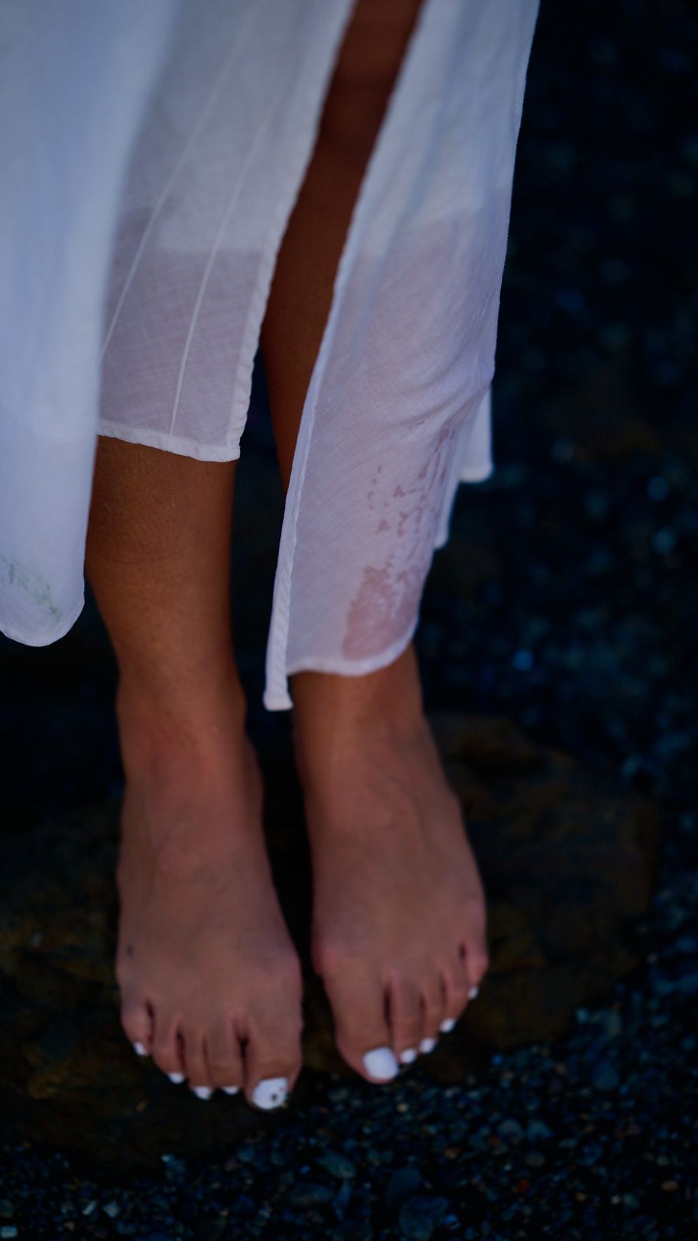 Bare feet with white painted toenails peek out from beneath a flowing white dress against a dark background.
