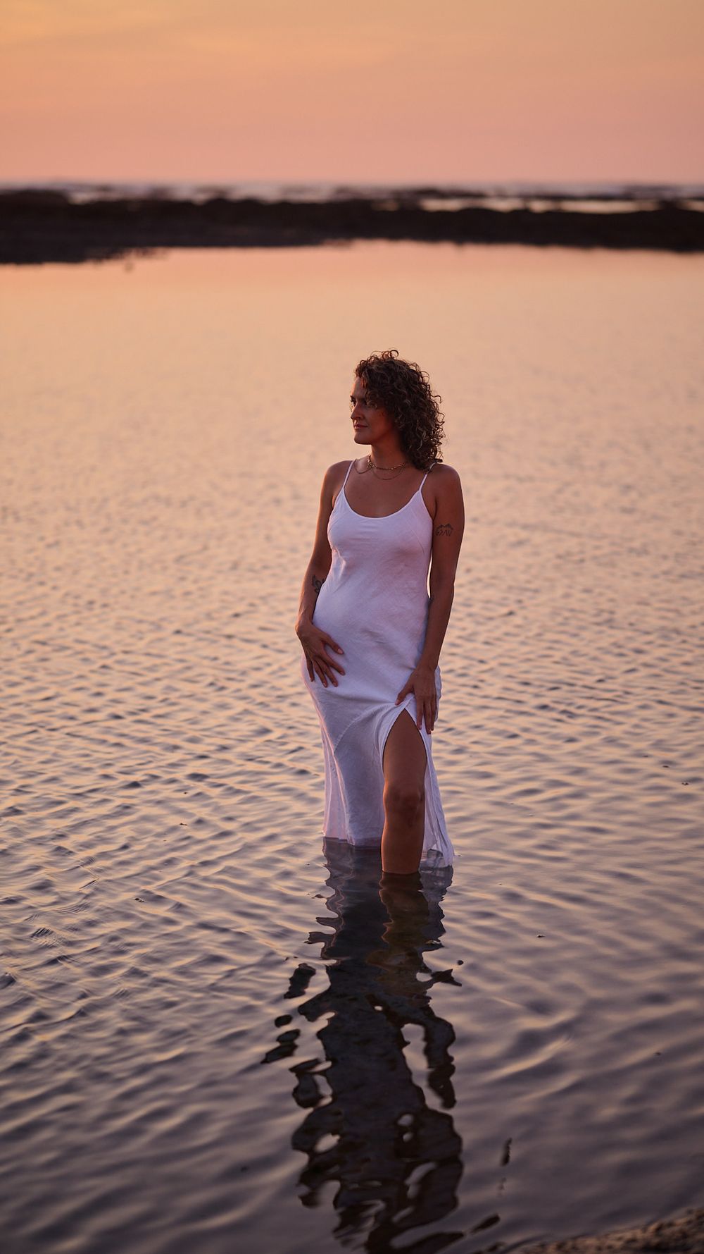 A person in a white dress stands in calm waters during sunset, creating a beautiful silhouette reflection.