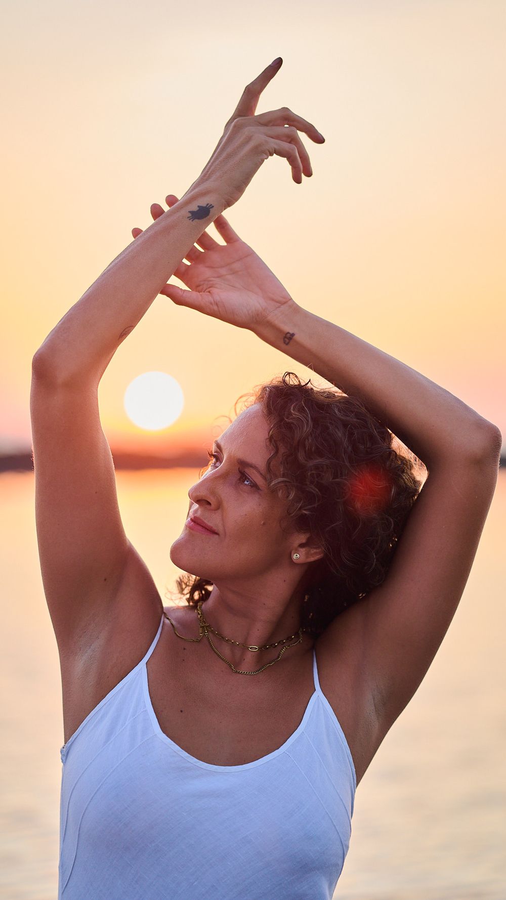 A dancer in a white tank top performs graceful arm movements against a golden sunset over water.