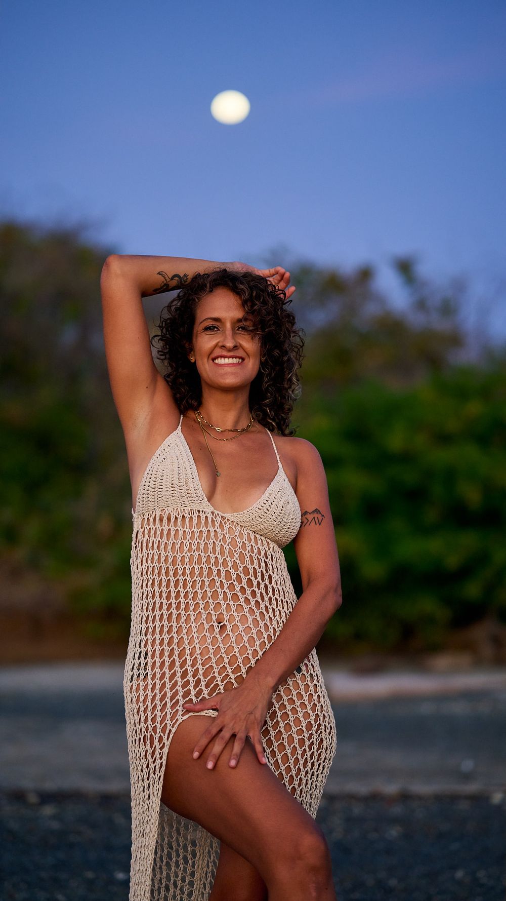 Woman in crochet beach cover up dress poses outdoors at dusk with full moon in background.