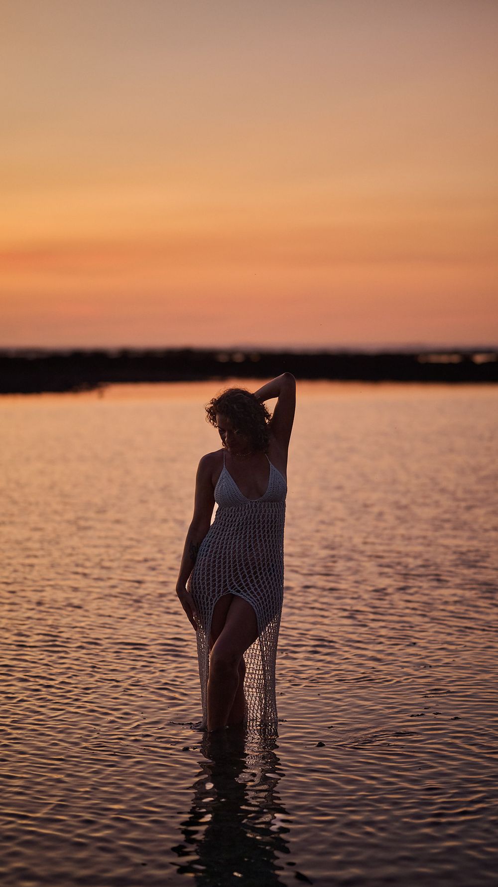 Silhouette of someone in a dress standing in calm water during a golden sunset.