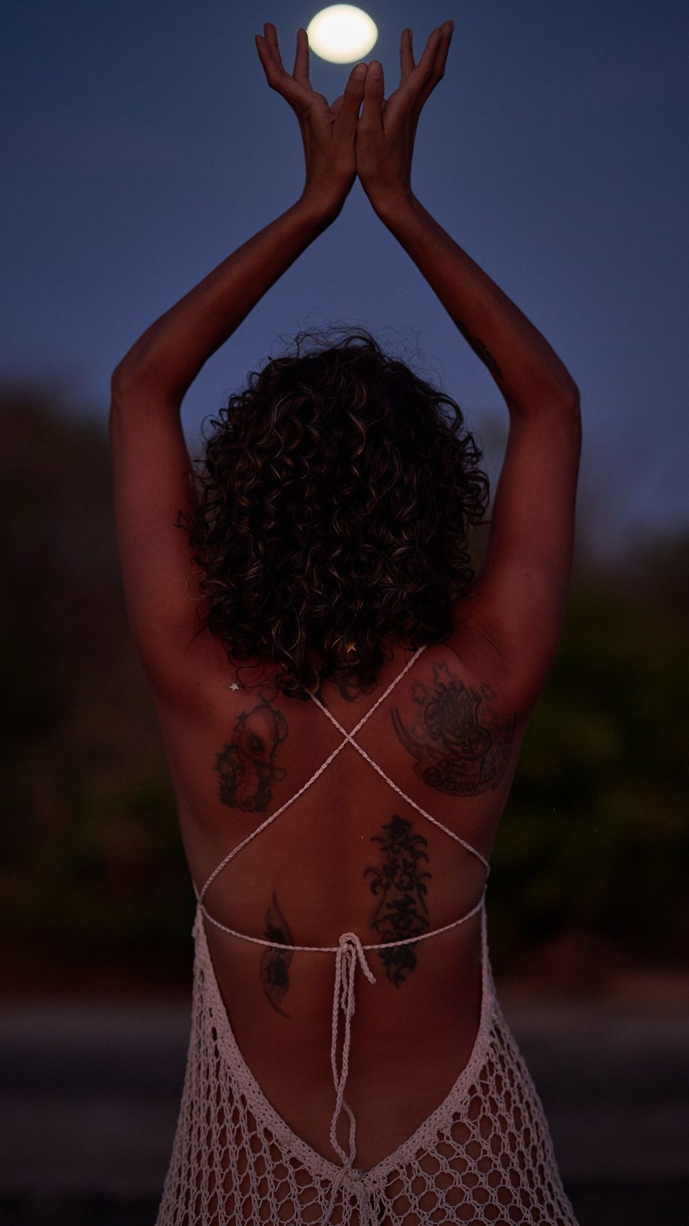 An artistic silhouette shows raised arms holding the moon in a mystical nighttime scene with tattoos and a crocheted top.