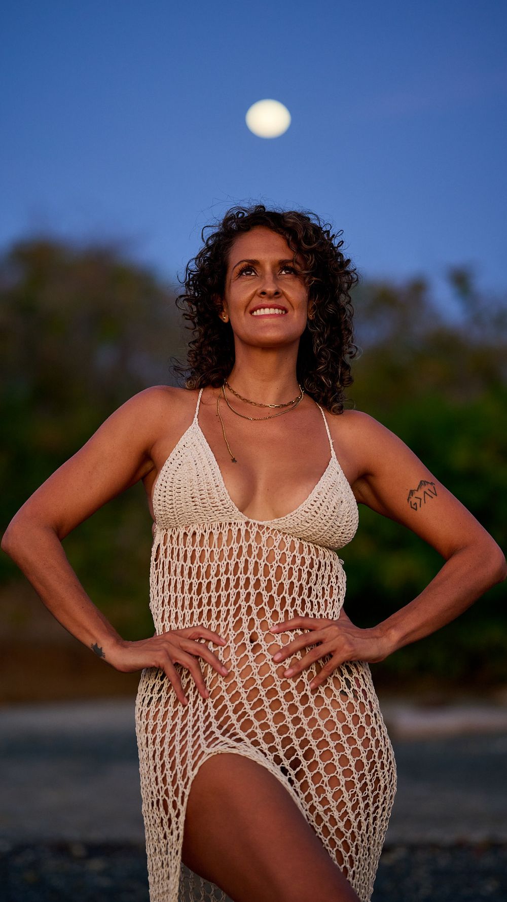 Someone poses in a crocheted white beach dress at sunset with a full moon in the blue sky.
