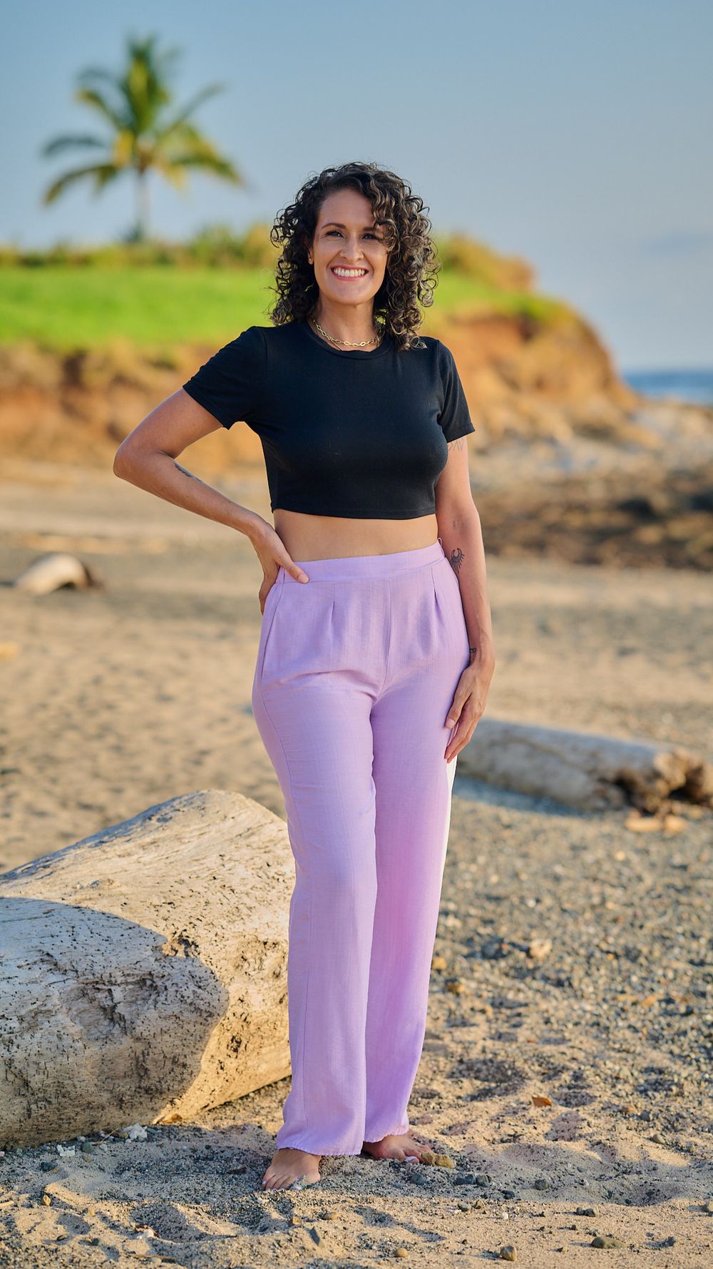 A casual beach outfit with a black crop top and lilac pants is modeled on a scenic Hawaiian beach with palm trees.