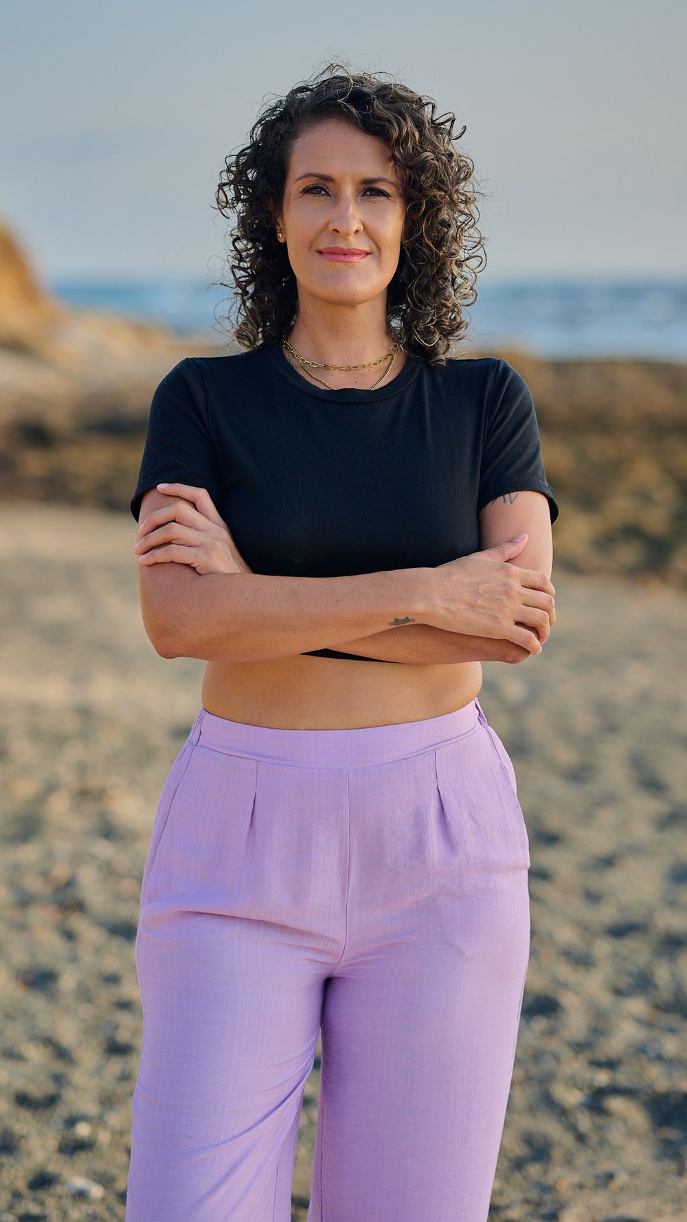 Person wearing a black top and purple pants standing with arms crossed on a beach at sunset.