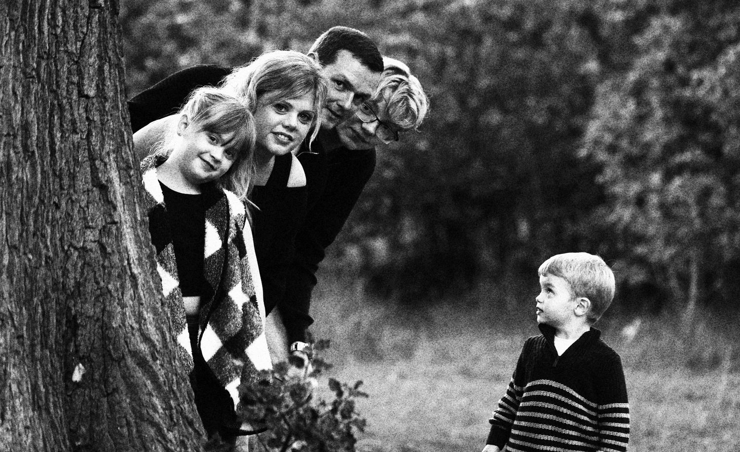 Black and white photo showing people in casual clothing sitting and leaning against a tree outdoors.