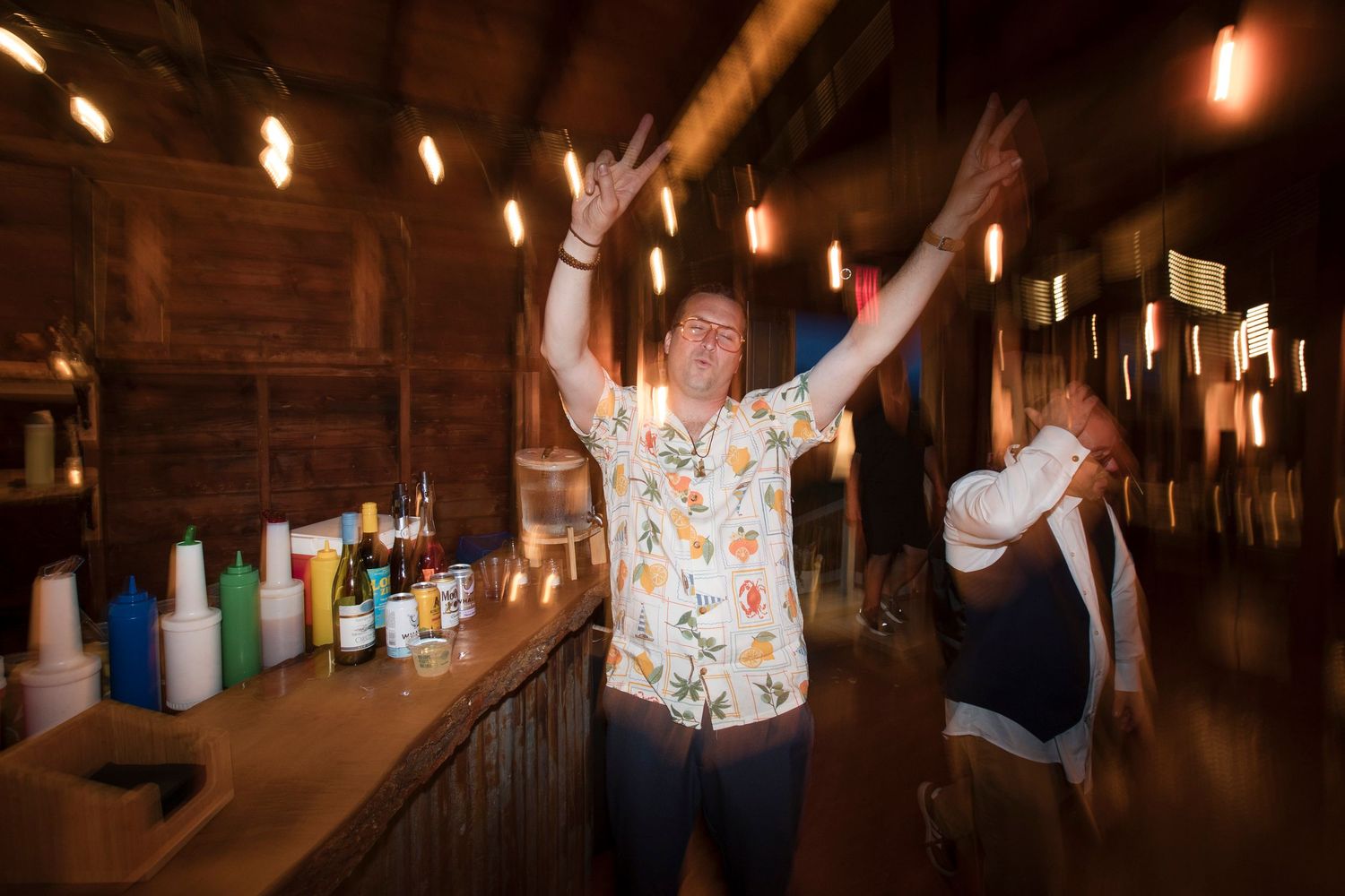 People dancing and celebrating inside a dimly lit rustic bar with glowing lights and bottles on the counter.