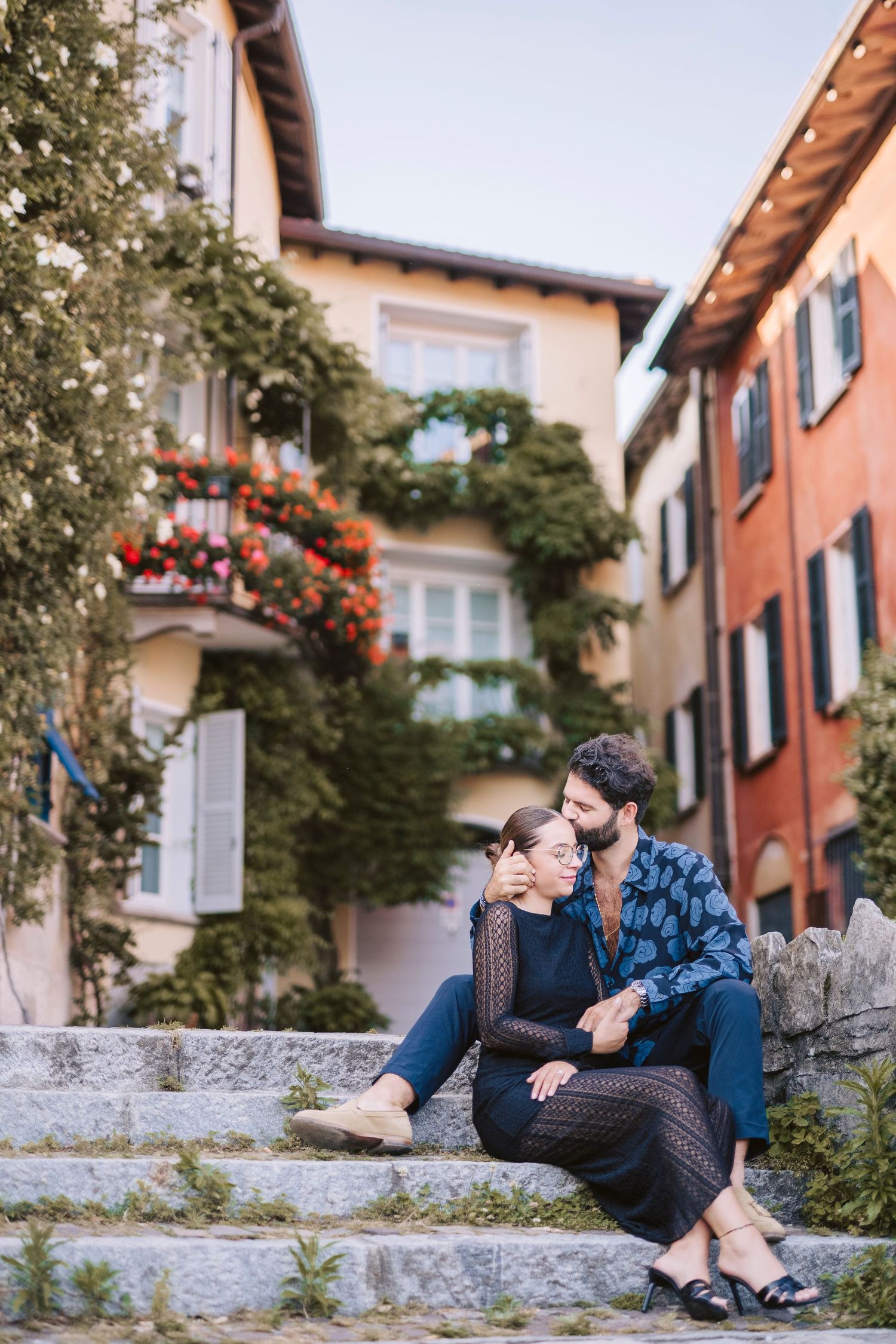 Giovane coppia siede sui gradini di un piccolo paese e si scambia un bacio affettuoso. Sullo sfondo case colorate e rampicanti fioriti.