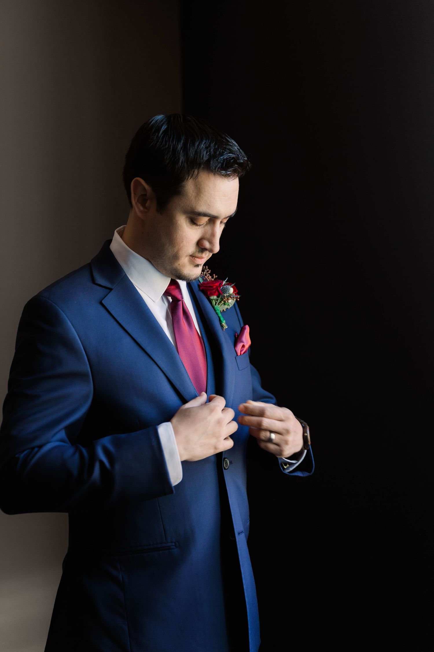 Groom in navy suit adjusting his boutonniere in dramatic lighting.