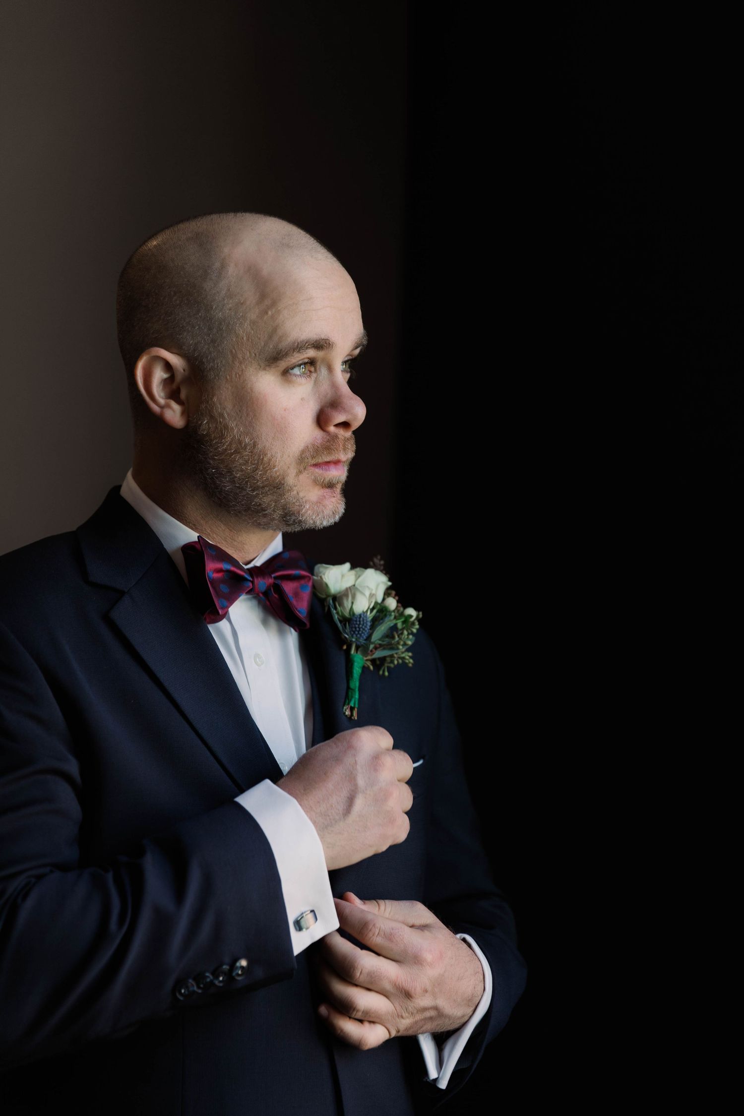 Close-up portrait of groom in navy suit adjusting boutonniere in dramatic lighting.