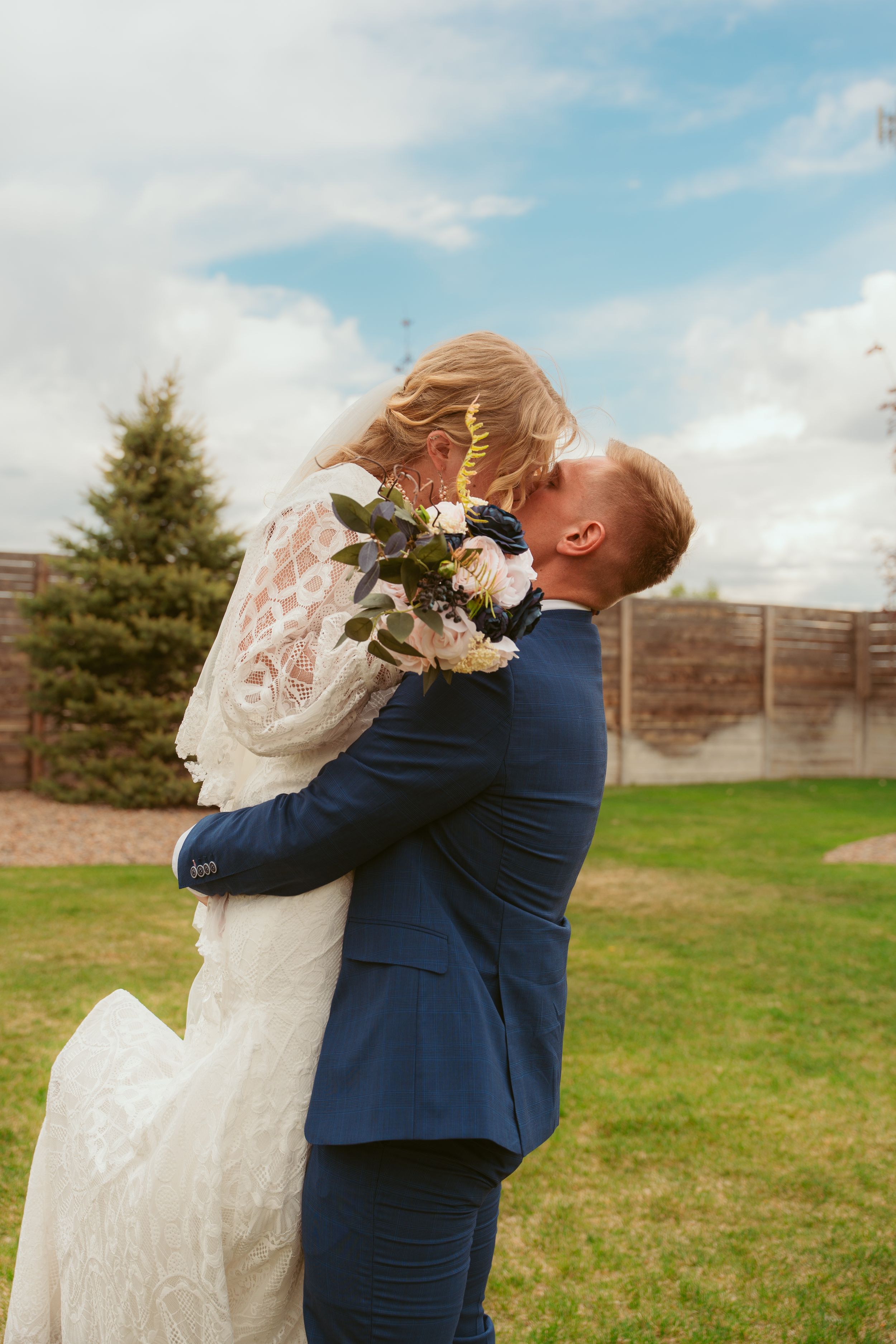 Joyful wedding moment captured as newlyweds embrace outdoors with the bride holding a bouquet against a wooden fence.