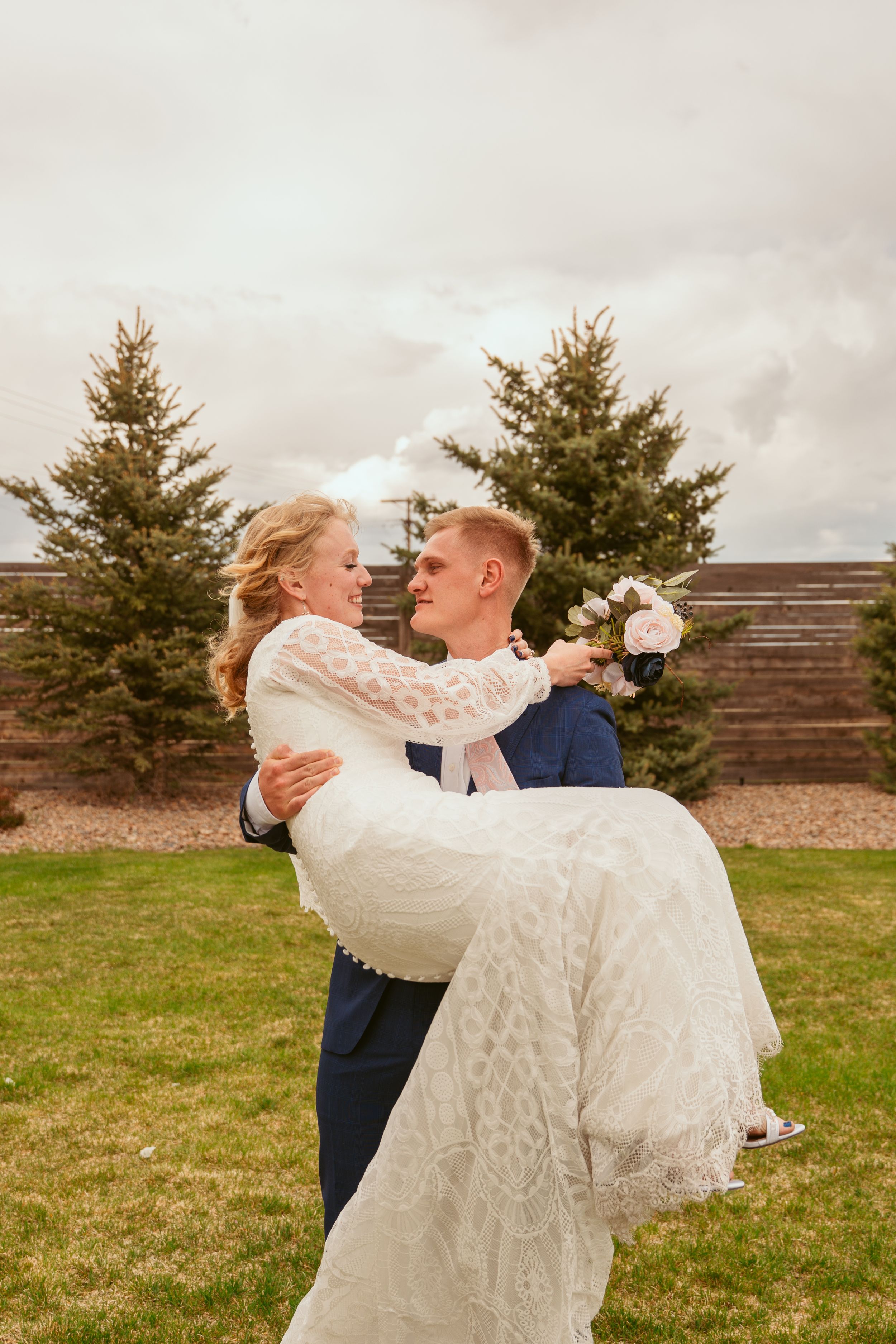 A romantic wedding portrait of a groom in navy lifting his bride in a lace dress against evergreen trees.