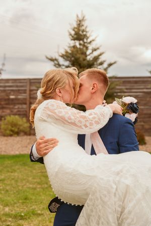 A romantic wedding moment captured outdoors as a couple in formal attire share a tender kiss while the groom carries the bride.
