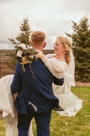 A couple in wedding attire dances joyfully together on a grassy field with evergreen trees in the background.
