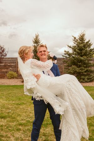 A joyful wedding moment captured as groom carries bride in white lace dress against an outdoor backdrop with evergreens.