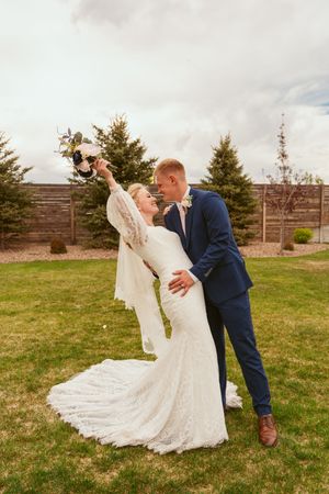 A romantic outdoor wedding photo with newlyweds embracing in a lush garden setting with evergreen trees in the background.