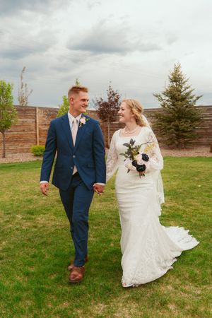 Newlyweds walk together holding hands in garden setting with wooden fence backdrop.