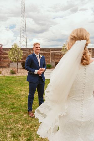 A first look wedding moment captured outdoors with a flowing white veil and navy blue suit against a cloudy sky.
