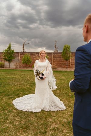 A wedding couple poses in a backyard garden with cloudy skies, the bride wearing an elegant lace gown with a long train.