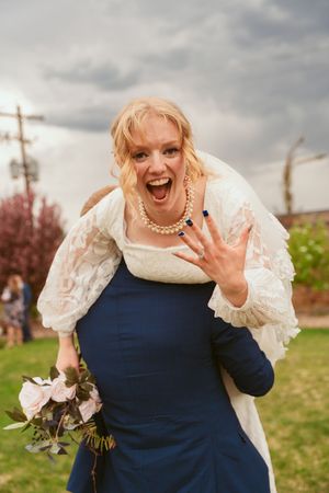 Joyful bride in lace dress shows off ring while holding bouquet outdoors.