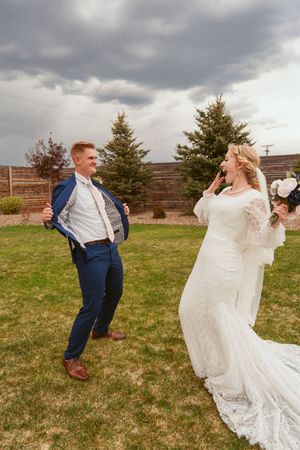 Dancing couple in wedding attire spins playfully on a grassy field under dramatic cloudy skies.