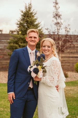 Newlywed couple in blue suit and lace wedding dress pose with bouquet featuring blue and white flowers in outdoor setting.