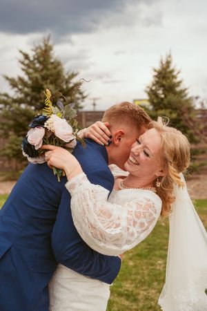 Joyful wedding couple embraces outdoors with bride holding bouquet against evergreen trees and cloudy sky.
