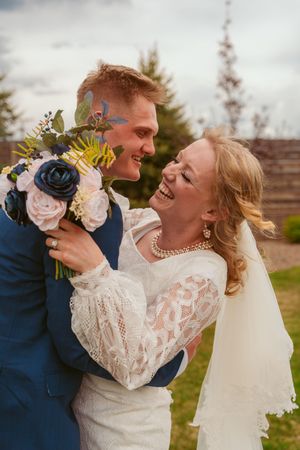 A joyful wedding couple shares a laugh while holding a bouquet with blue and pink flowers against an outdoor backdrop.