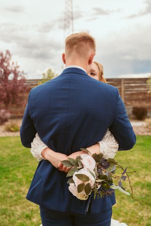 Back view of couple embracing at outdoor wedding with bride holding rustic bouquet against cloudy sky.