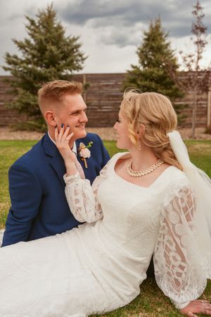 A romantic wedding portrait of a couple outdoors with the bride in lace sleeves and groom in navy suit against pine trees.