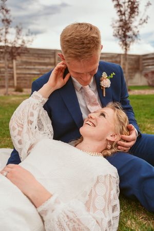 A playful wedding photo of a couple lying in grass, with the bride in lace and groom in navy sharing an intimate moment.
