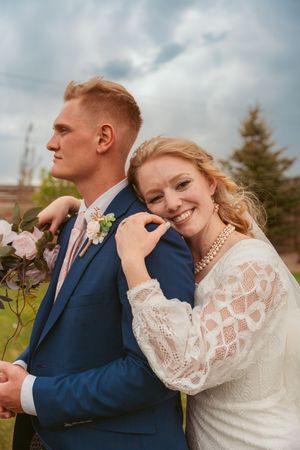 A romantic wedding photo shows a couple embracing outdoors, with the bride in lace and groom in navy against moody clouds.