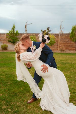 A romantic wedding dip pose on a lawn with evergreen trees and a wooden fence in the background.