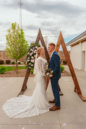 Wedding couple poses in front of wooden A-frame arch decorated with white floral arrangements.