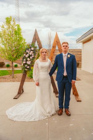 Bride in lace gown and groom in navy suit pose in front of rustic wooden decorative panels.