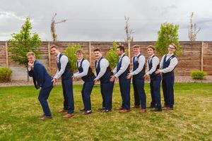A playful line of groomsmen in navy vests and slacks lean forward in a cascading domino effect pose on grass.