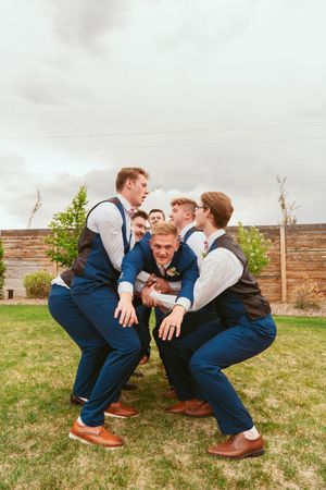 Groomsmen in navy vests playfully pose and crouch together on grass during wedding photoshoot.