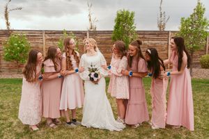 Group photo of bridesmaids in blush pink dresses standing with bride in white gown against wooden fence backdrop.