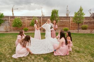 Wedding party arranges the long train of a white lace wedding dress on a lawn against a wooden fence.